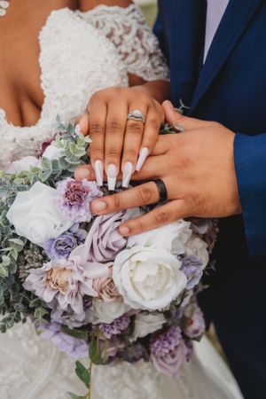 Wedding detail shot of hands with rings over a beautiful purple and white flower bouquet.
