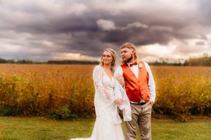 Wedding couple poses in autumn field with stormy clouds, groom wearing orange vest.