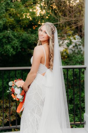 A bride looks over her shoulder while holding a coral bouquet on a white deck.