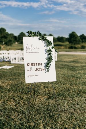 Elegant wedding welcome sign decorated with greenery displayed on grass with white chairs in background.
