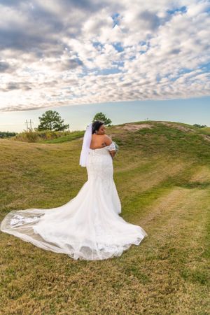 A bride in a fitted lace wedding gown with flowing train poses on a grassy hilltop under dramatic cloudy skies.