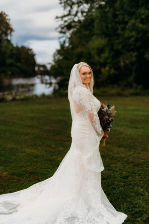 Elegant bride in long-sleeved lace wedding gown poses outdoors with bouquet against natural backdrop.