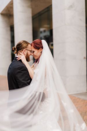 Intimate moment between newlyweds as veil flows in gentle breeze by stone columns.