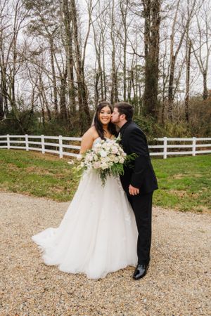 A romantic wedding portrait of a couple standing on a gravel path with a white fence and wooded background.