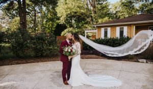 A wedding couple poses outdoors while bride's long lace veil flows dramatically in the wind.