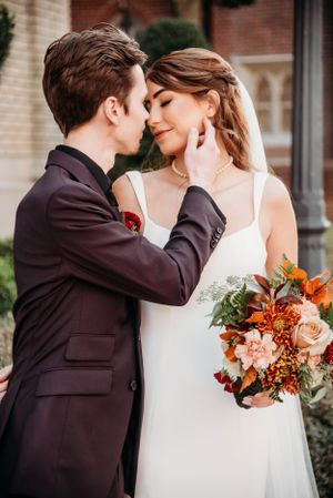 Intimate wedding portrait with burgundy suit and autumn bouquet against brick building.