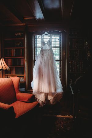 A white lace wedding dress hangs in a dark, elegant library room with rich wood details.