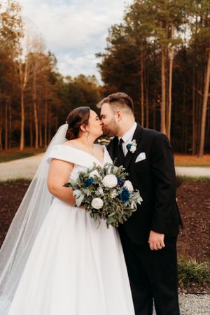 A romantic wedding kiss with a blue and white bouquet against an autumn forest backdrop.