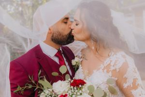 A romantic wedding moment with couple kissing under flowing veil, groom in burgundy suit.