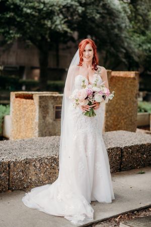 Elegant bride with red hair poses with floral bouquet in white lace wedding gown.