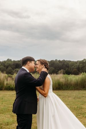 Newlyweds share intimate moment in outdoor field setting under cloudy sky.