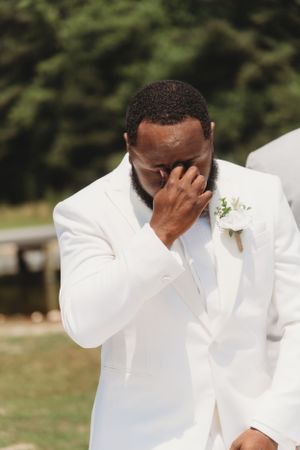 Groom in white tuxedo shows emotional reaction during wedding ceremony with delicate floral boutonniere.