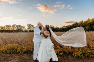 A wedding couple poses in a field at sunset as the bride's veil flows in the wind.