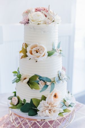 Elegant three-tier white wedding cake decorated with blush roses and green foliage on a crystal cake stand.