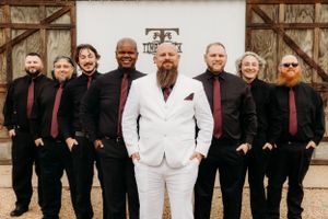 Wedding party poses with groomsmen in black suits and groom in white suit.
