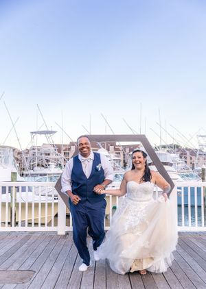 Newlyweds running joyfully on marina boardwalk with boats in background.