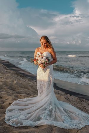 Bride in lace gown with flowing train poses on beach with floral bouquet.