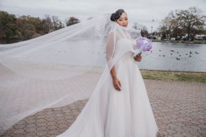 Long flowing veil catches the breeze as bride in white gown holds purple flowers by waterfront.