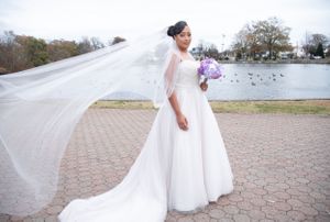 Elegant wedding dress with flowing veil and purple bouquet captured by a lake on a cloudy day.