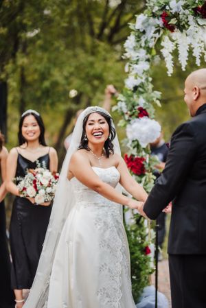 Joyful wedding ceremony moment under floral arch with bridesmaids in black dresses.