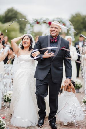 Happy newlyweds exit ceremony under floral arch with flower girl in matching white dress.