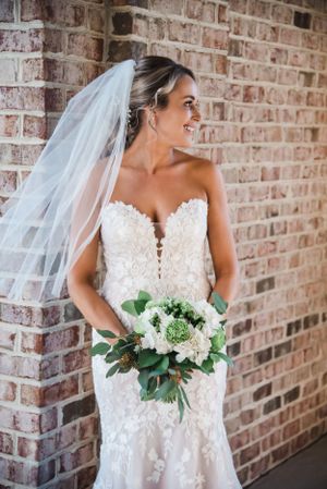 Radiant bride in lace wedding dress holds white bouquet against brick wall.