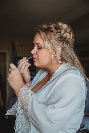 A person in a white robe applies makeup while looking in a mirror during wedding preparations.