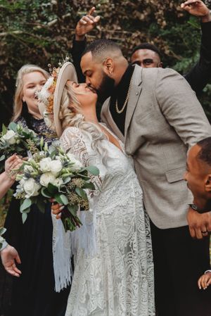 Romantic wedding kiss captured outdoors with wedding party celebrating in the background.