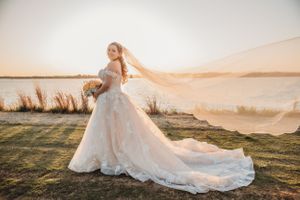 Bride in flowing dress with veil catching wind at sunset by waterfront.