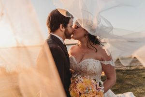 Romantic sunset wedding kiss with veil catching wind and yellow bouquet.