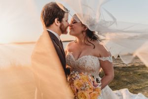 Ethereal wedding moment captured at sunset with flowing veil and yellow bouquet against nature backdrop.