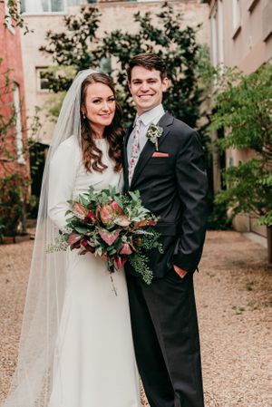 Formal wedding portrait in garden courtyard features classic attire and lush floral bouquet.