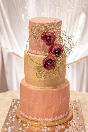Three-tier wedding cake with blush and gold tones adorned with burgundy sugar flowers.