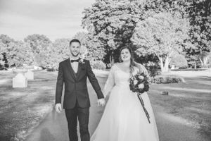 A couple walks hand in hand down a tree-lined path in formal wedding attire.