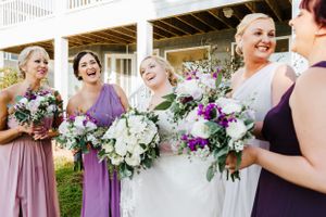 A joyful wedding party holding bouquets in purple and white dresses on a porch.