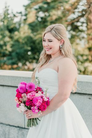 A bride in a strapless white gown holds a bright pink bouquet while standing outdoors.