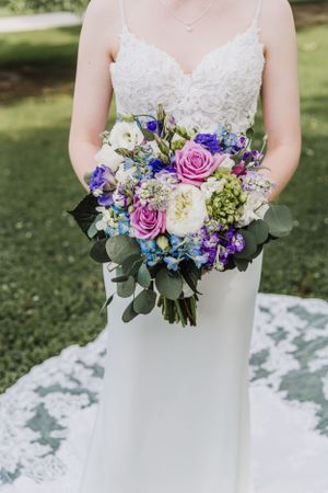 Elegant bridal bouquet featuring pink roses, purple flowers, and eucalyptus held against white wedding dress.