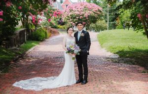 Beautiful wedding couple poses on brick pathway surrounded by blooming pink flowers.
