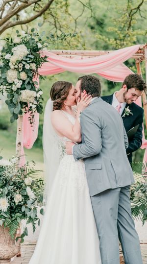 Newlyweds share their first kiss under a pink draped arch decorated with white flowers.