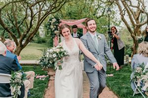 A joyful wedding couple walks down an outdoor aisle after their ceremony surrounded by greenery and guests.