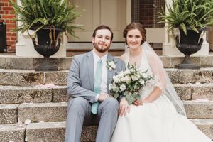 A wedding couple sits on stone steps decorated with potted plants at a historic building entrance.