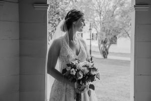 A black and white portrait of a bride holding a bouquet while standing under a columned portico.
