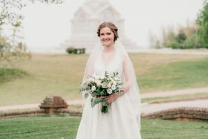 A bride in a flowing white dress and veil holds a bouquet of greenery and flowers in an outdoor setting.