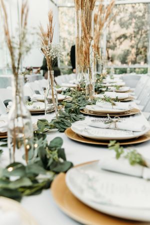 Elegant table setting with gold chargers, white plates, eucalyptus garland and dried pampas grass centerpieces.