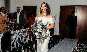 A bride walks down an aisle carrying a purple bouquet with guests in background.