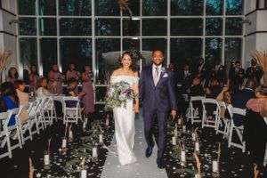A couple walks down an aisle decorated with candles and white chairs.