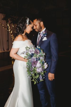 A couple in wedding attire share an intimate moment with a purple floral bouquet.