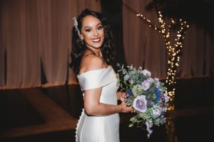 A bride in an off-shoulder white gown holds a purple and blue bouquet while smiling.