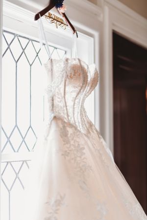 Elegant white lace wedding dress hangs in window with diamond pattern leaded glass.
