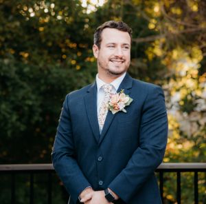 Groom in navy suit with floral boutonniere smiles during outdoor wedding.